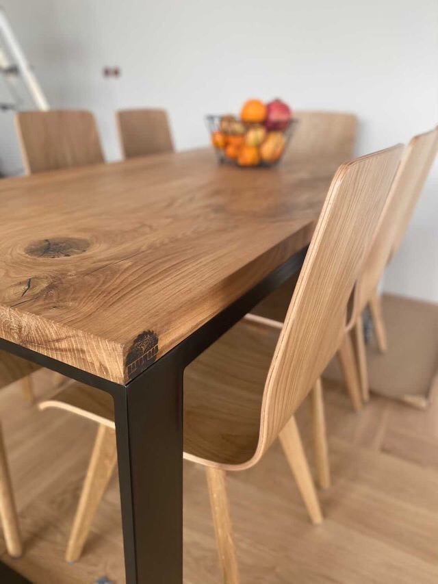 Stół z blatem z litego drewna z widocznym sękiem i czarnymi metalowymi nogami, otoczony jasnymi, drewnianymi krzesłami o zaokrąglonym oparciu; na stole koszyk z owocami.