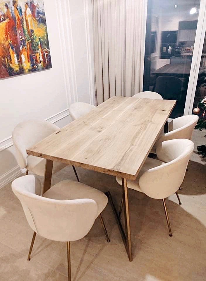 Stół z blatem z jasnego drewna o wyraźnym usłojeniu, wsparty na złotych, skośnych nogach, otoczony przez sześć kremowych krzeseł z welurową tapicerką i złotymi nogami, w tle obraz i zasłony.