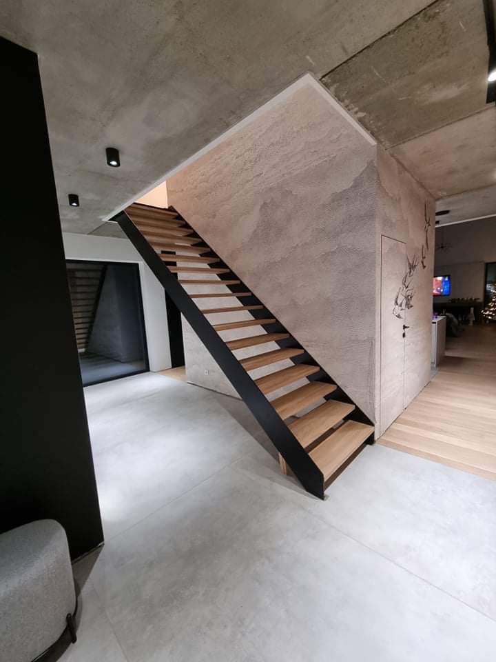 Nowoczesne schody z drewnianymi stopniami i czarną metalową konstrukcją w minimalistycznym wnętrzu z betonowym sufitem i ścianami o surowym wykończeniu.