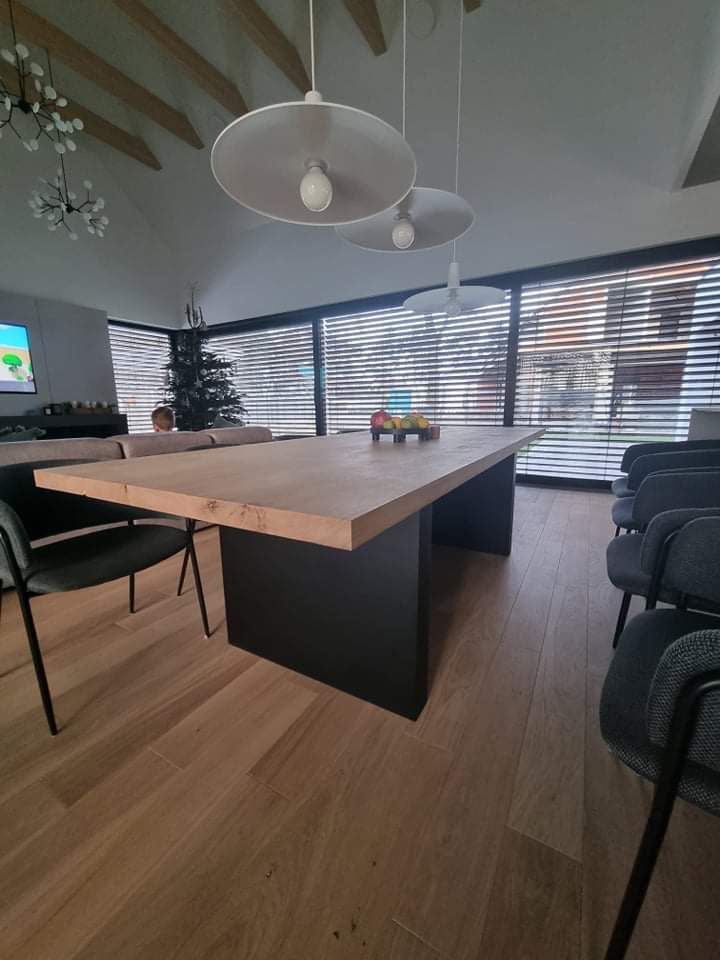 Stół z blatem z jasnego drewna i czarną, masywną podstawą w nowoczesnym wnętrzu z dużymi oknami i drewnianą podłogą.