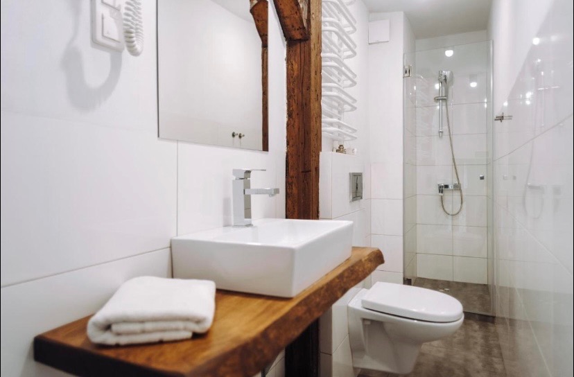 Nowoczesna łazienka z umywalką nablatową na blacie z surowego drewna, widoczna kolumna z drewna konstrukcyjnego, białe płytki i kabina prysznicowa w tle.