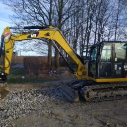 Żółta koparka gąsienicowa CAT 308E z łyżką nabierającą żwir na tle drzew bez liści i błękitnego nieba.