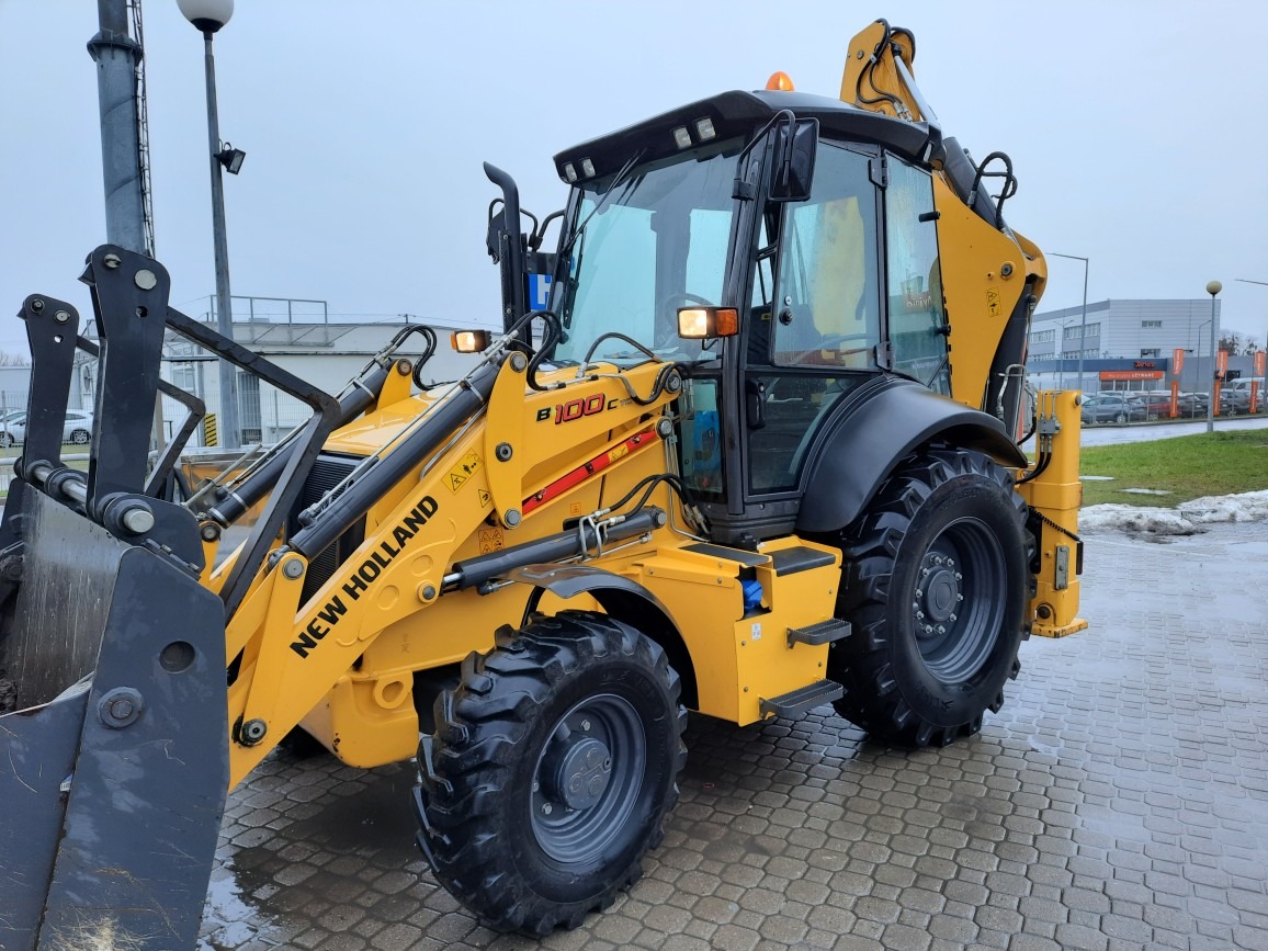 Żółta koparko-ładowarka New Holland B100C na kostce brukowej w pochmurny dzień, z widocznymi śladami błota na oponach i łyżce.