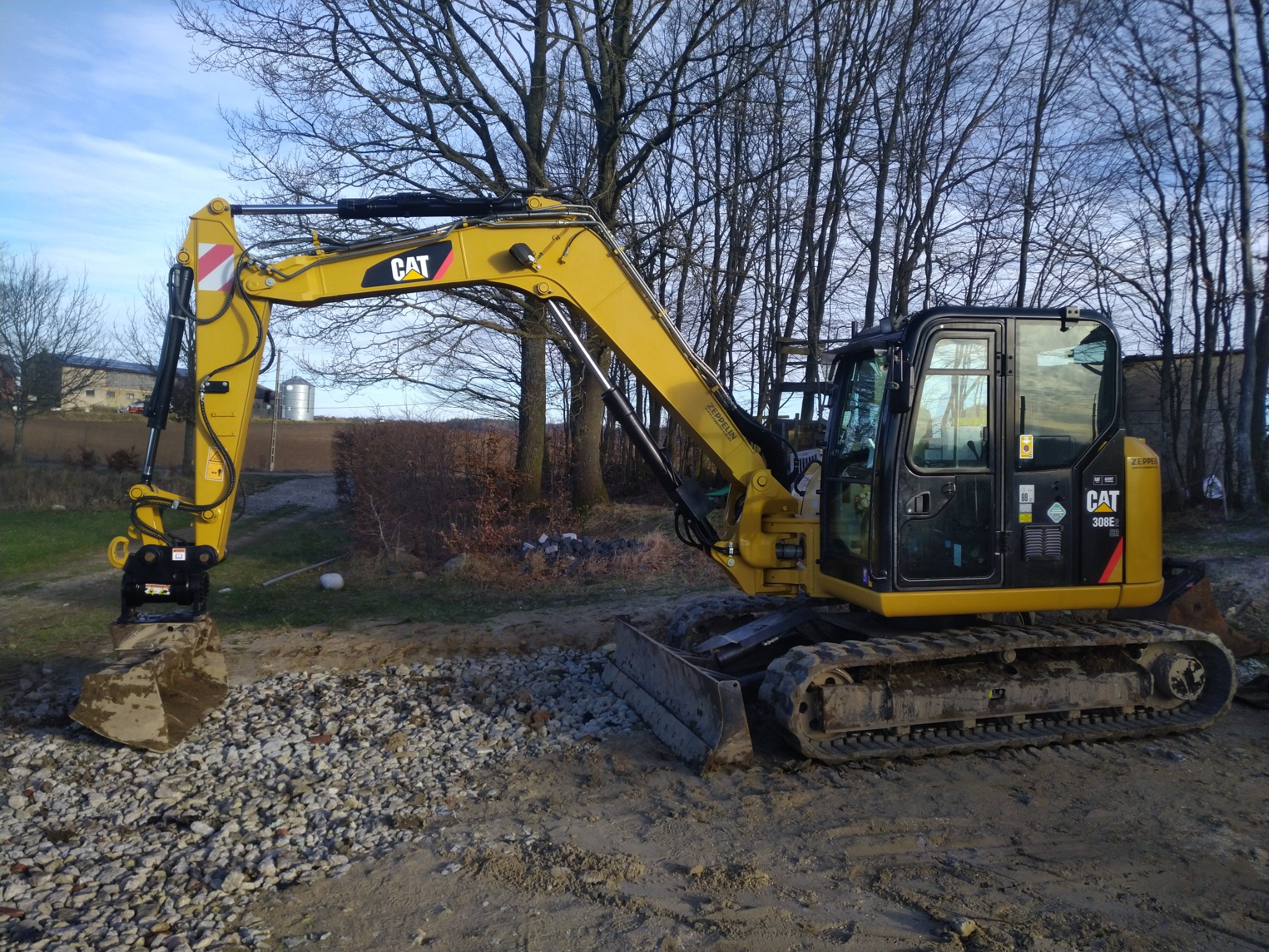 Żółta koparka gąsienicowa CAT 308E z łyżką nabierającą żwir na tle drzew bez liści i błękitnego nieba.