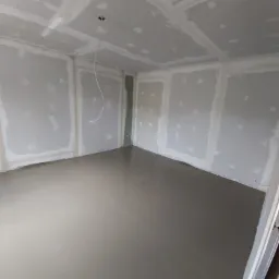 Surowe wnętrze pokoju z wylaną, gładką posadzką, ścianami pokrytymi płytami kartonowo-gipsowymi przed szpachlowaniem, widoczny kabel zwisający z sufitu.