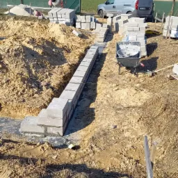 Wykopany fundament z ułożonymi bloczkami betonowymi tworzącymi ścianę, widoczna betoniarka i samochód dostawczy w tle.