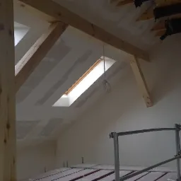 Poddasze w trakcie prac wykończeniowych, widoczne drewniane belki konstrukcyjne, okna dachowe i częściowo wykończone ściany, rusztowanie w pomieszczeniu.