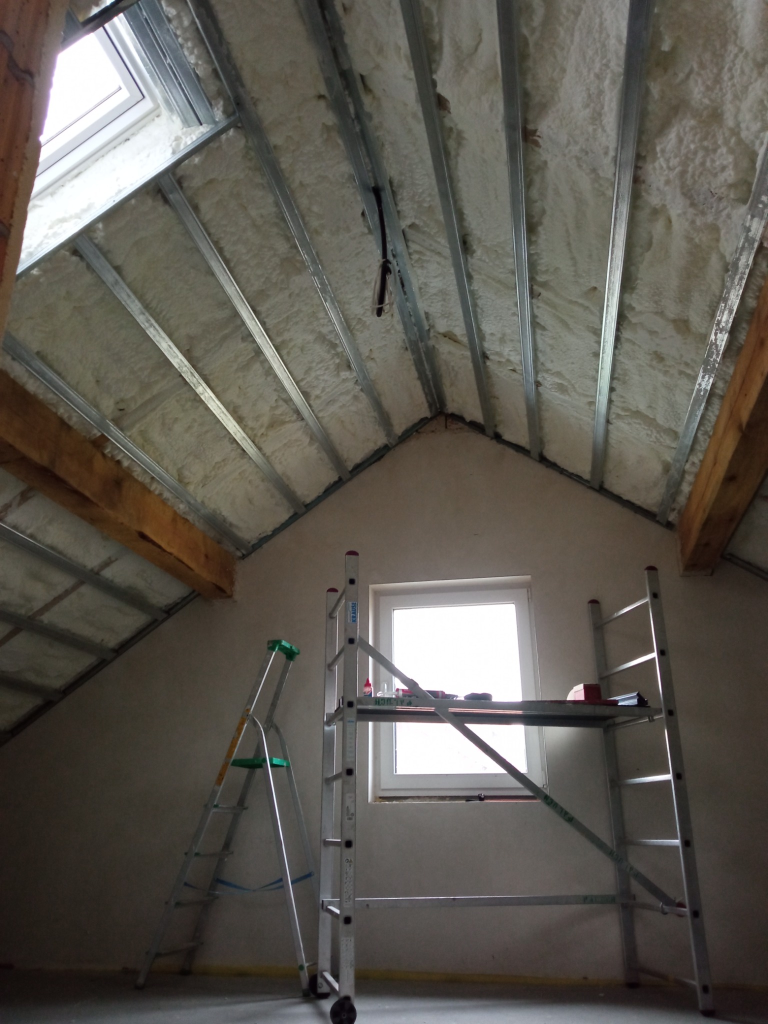 Poddasze w trakcie ocieplania pianką, widoczne metalowe profile pod płyty gipsowo-kartonowe, okno dachowe i drabiny.