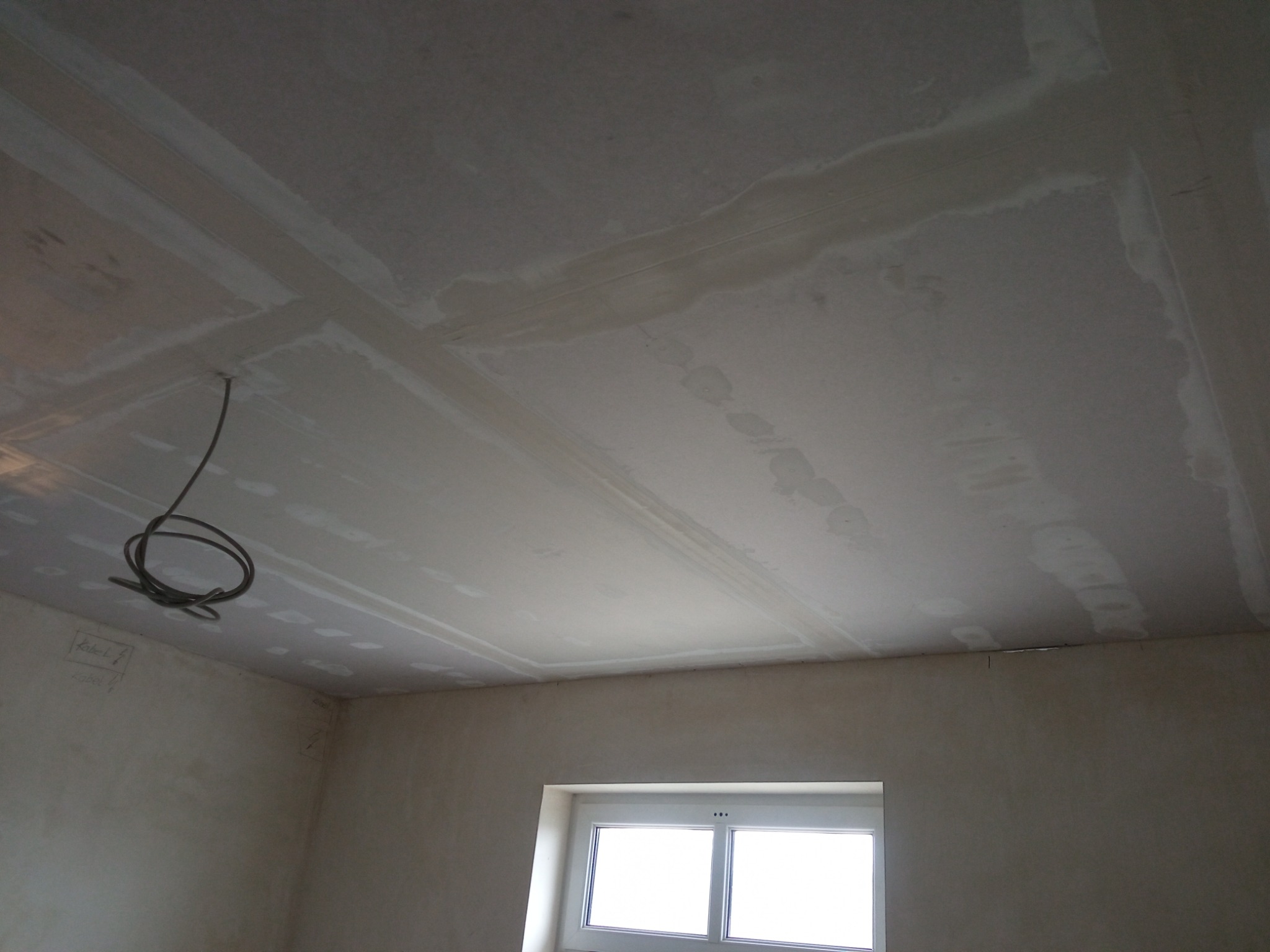 Płyty gipsowo-kartonowe na suficie w trakcie szpachlowania, widoczne łączenia i przewody elektryczne zwisające z sufitu, ściany przygotowane do malowania, okno w dolnej części kadru.
