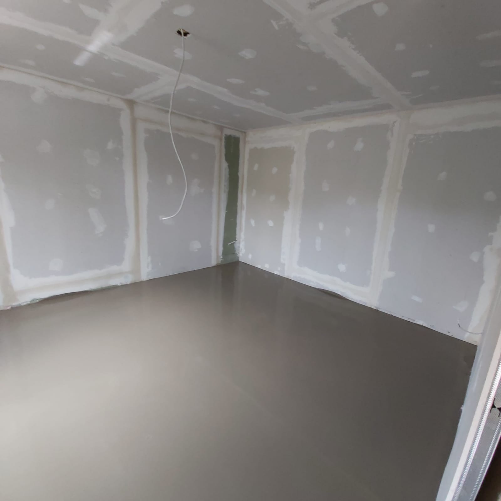 Surowe wnętrze pokoju z wylaną, gładką posadzką, ścianami pokrytymi płytami kartonowo-gipsowymi przed szpachlowaniem, widoczny kabel zwisający z sufitu.