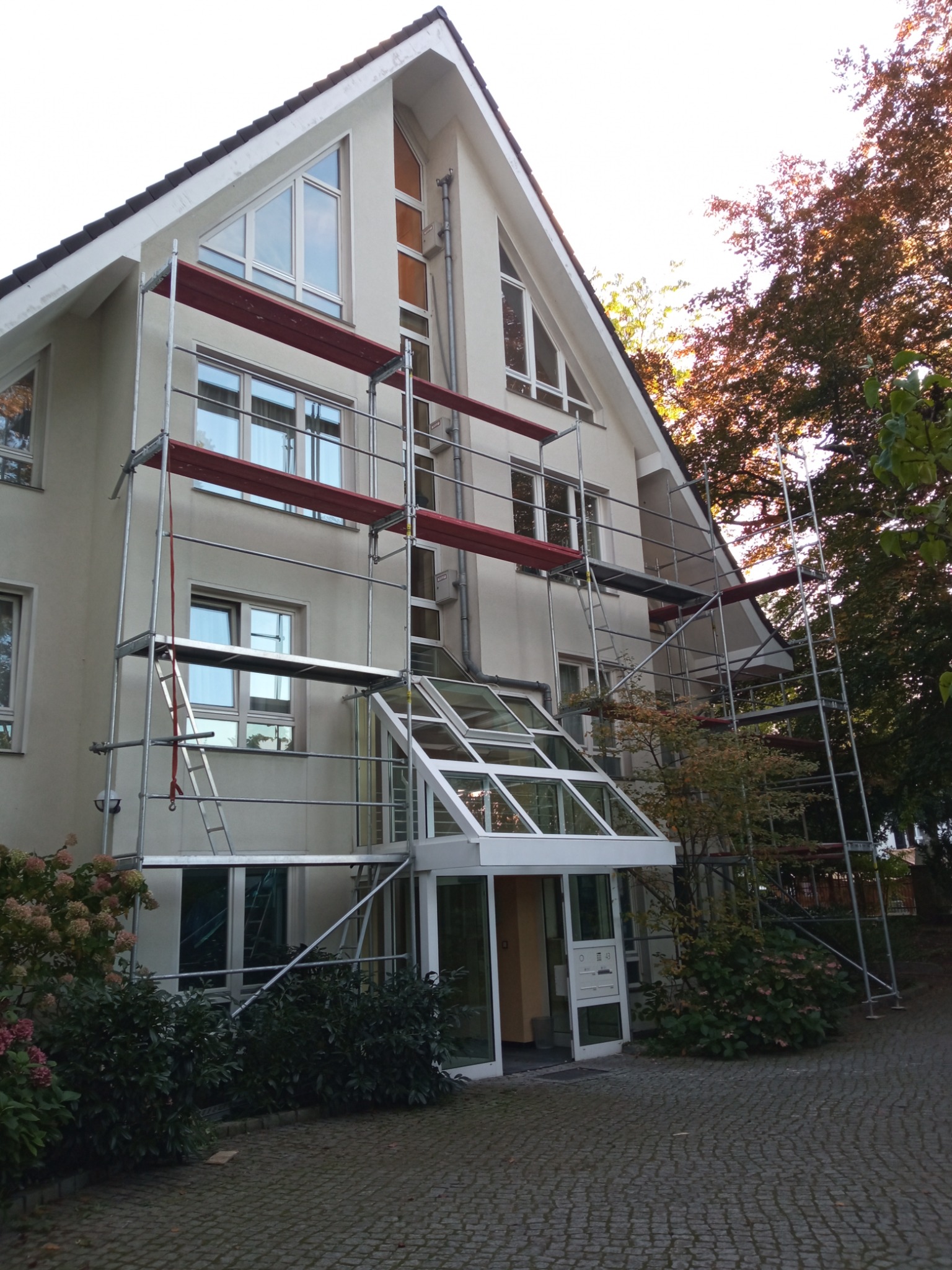 Dwupiętrowy budynek z jasną elewacją w trakcie prac remontowych, otoczony rusztowaniami i zielenią.