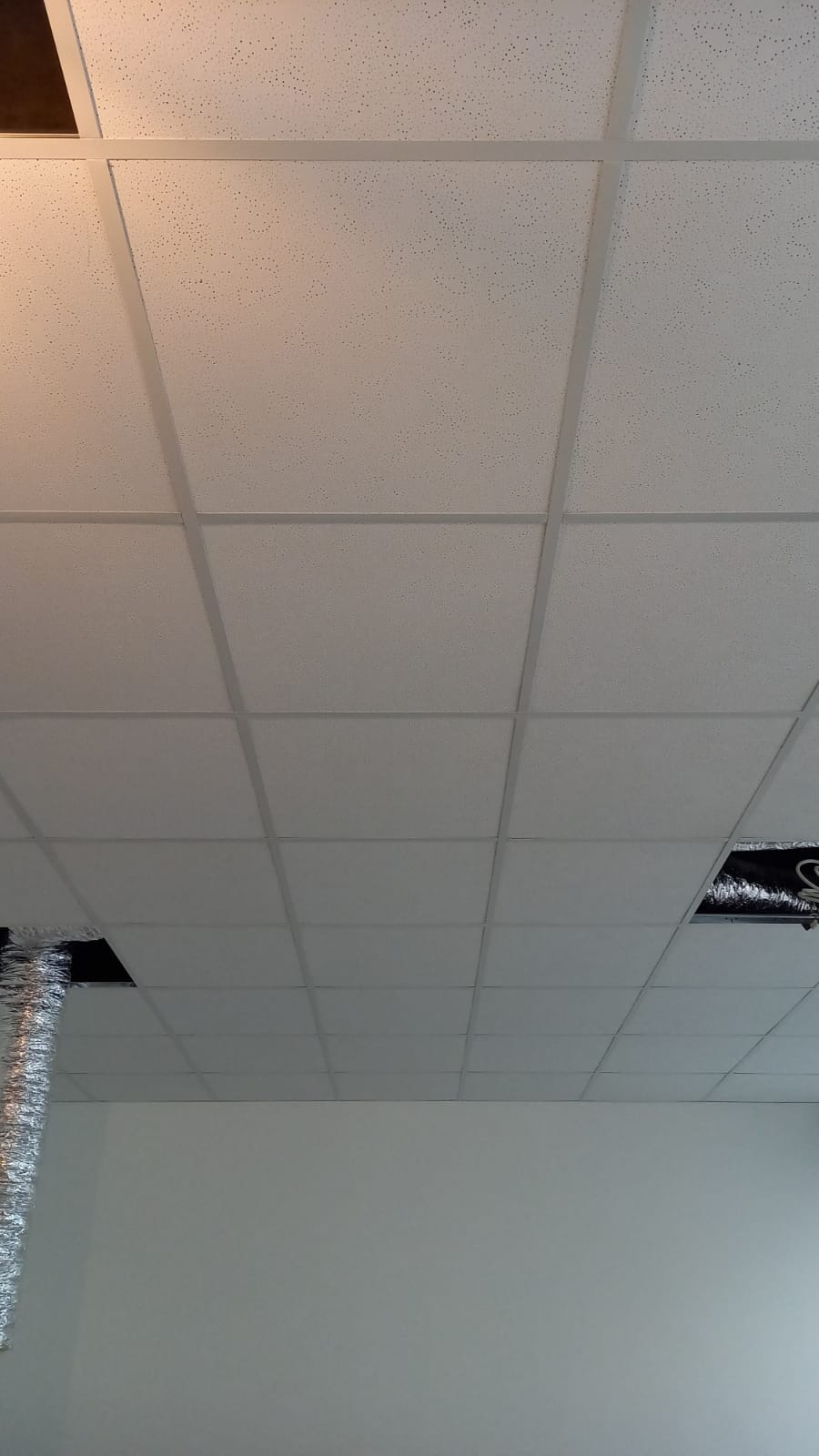 Sufit podwieszany z białych paneli rastrowych, widoczne otwory wentylacyjne i fragment instalacji w przestrzeni nad sufitem.