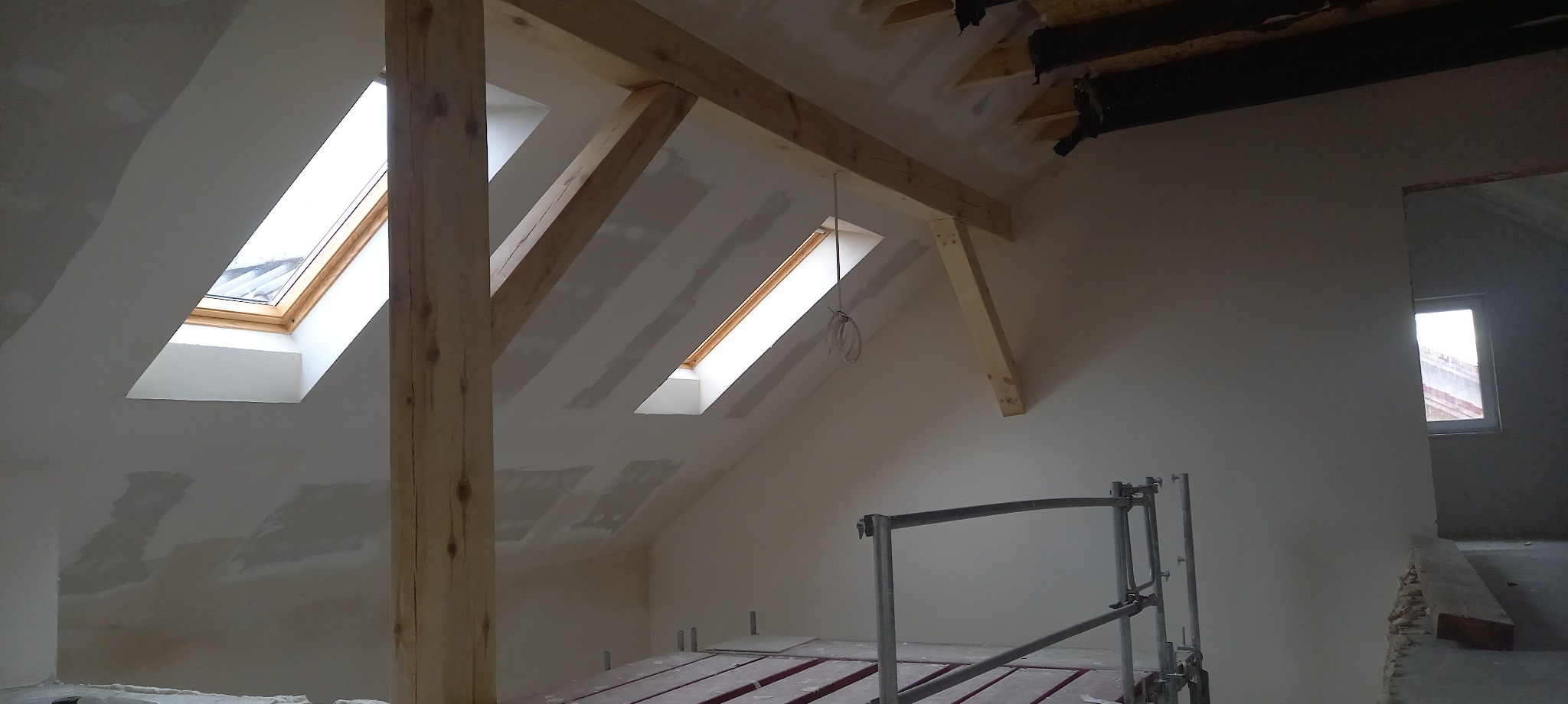 Poddasze w trakcie prac wykończeniowych, widoczne drewniane belki konstrukcyjne, okna dachowe i częściowo wykończone ściany, rusztowanie w pomieszczeniu.