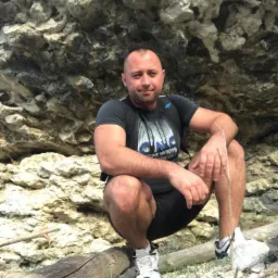 Mężczyzna w sportowym stroju kuca na pniu drzewa na tle skalnej ściany, uśmiechając się do kamery.