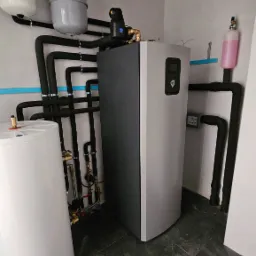 Szara pompa ciepła w kotłowni, widoczne czarne rury instalacji, zbiorniki wyrównawcze, manometry i zawory na tle szarej podłogi z płytek.