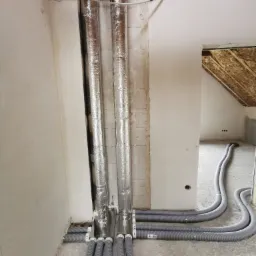 Montaż instalacji wentylacyjnej w surowym wnętrzu: rury spiro owinięte folią aluminiową pionowo, połączone z szarymi, karbowanymi przewodami poziomymi na podłodze. Widoczne surowe ściany...