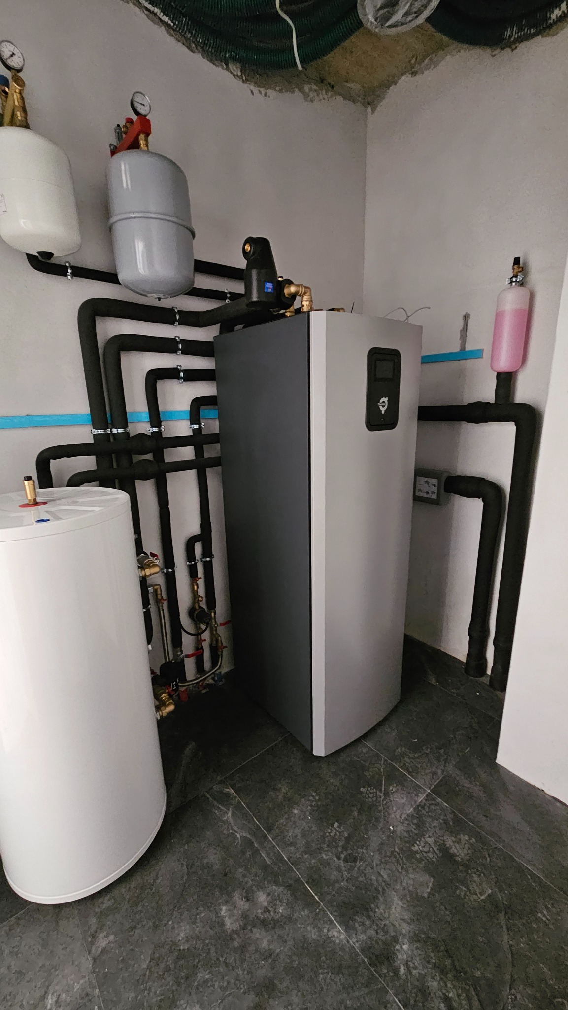 Szara pompa ciepła w kotłowni, widoczne czarne rury instalacji, zbiorniki wyrównawcze, manometry i zawory na tle szarej podłogi z płytek.