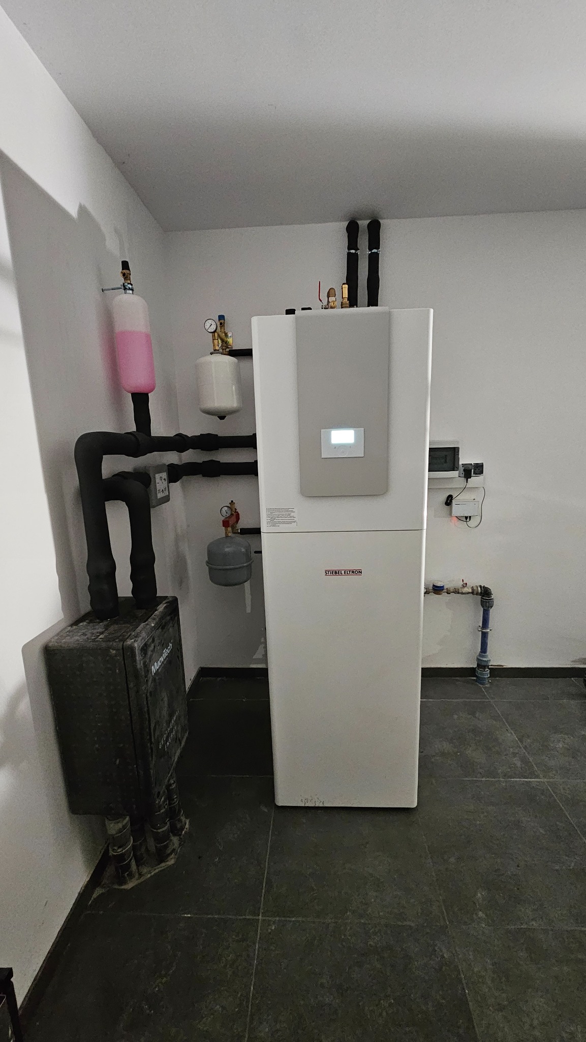 Biała pompa ciepła Stiebel Eltron z podłączonymi rurami, zbiornik wyrównawczy z różowym płynem, czarna skrzynka rozdzielcza Muovitech, szare płytki podłogowe w pomieszczeniu technicznym.