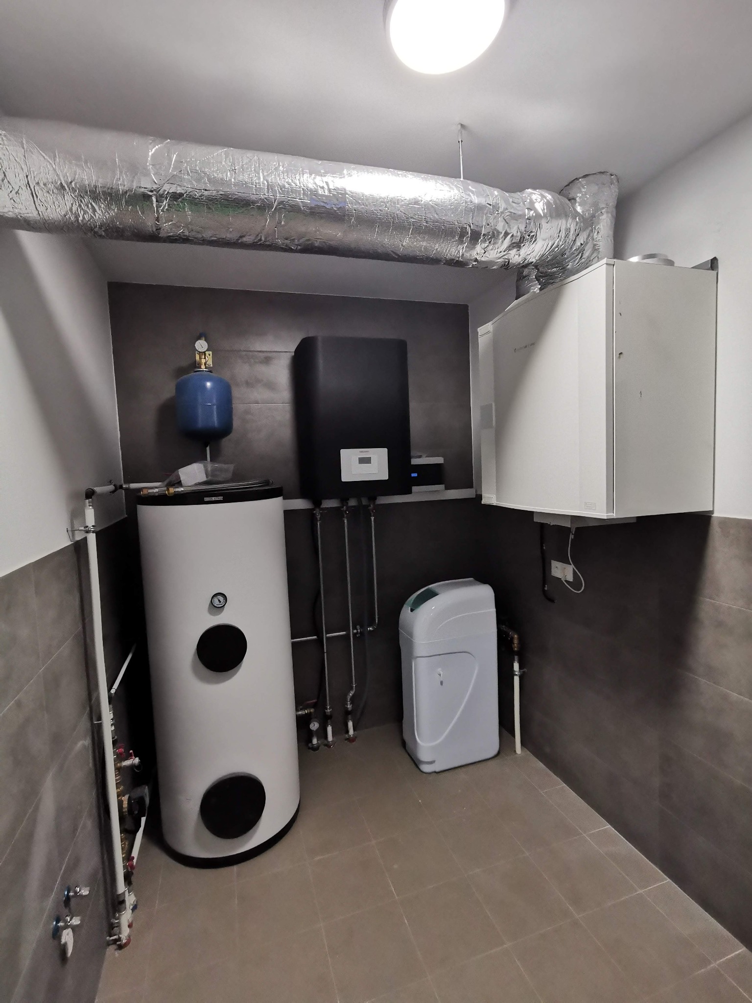 Kompletna kotłownia z zasobnikiem ciepłej wody, zmiękczaczem wody i rekuperatorem z wymiennikiem obrotowym System AIR VTR 300, widoczna instalacja rur i osprzętu na tle szarej ściany z płytek.