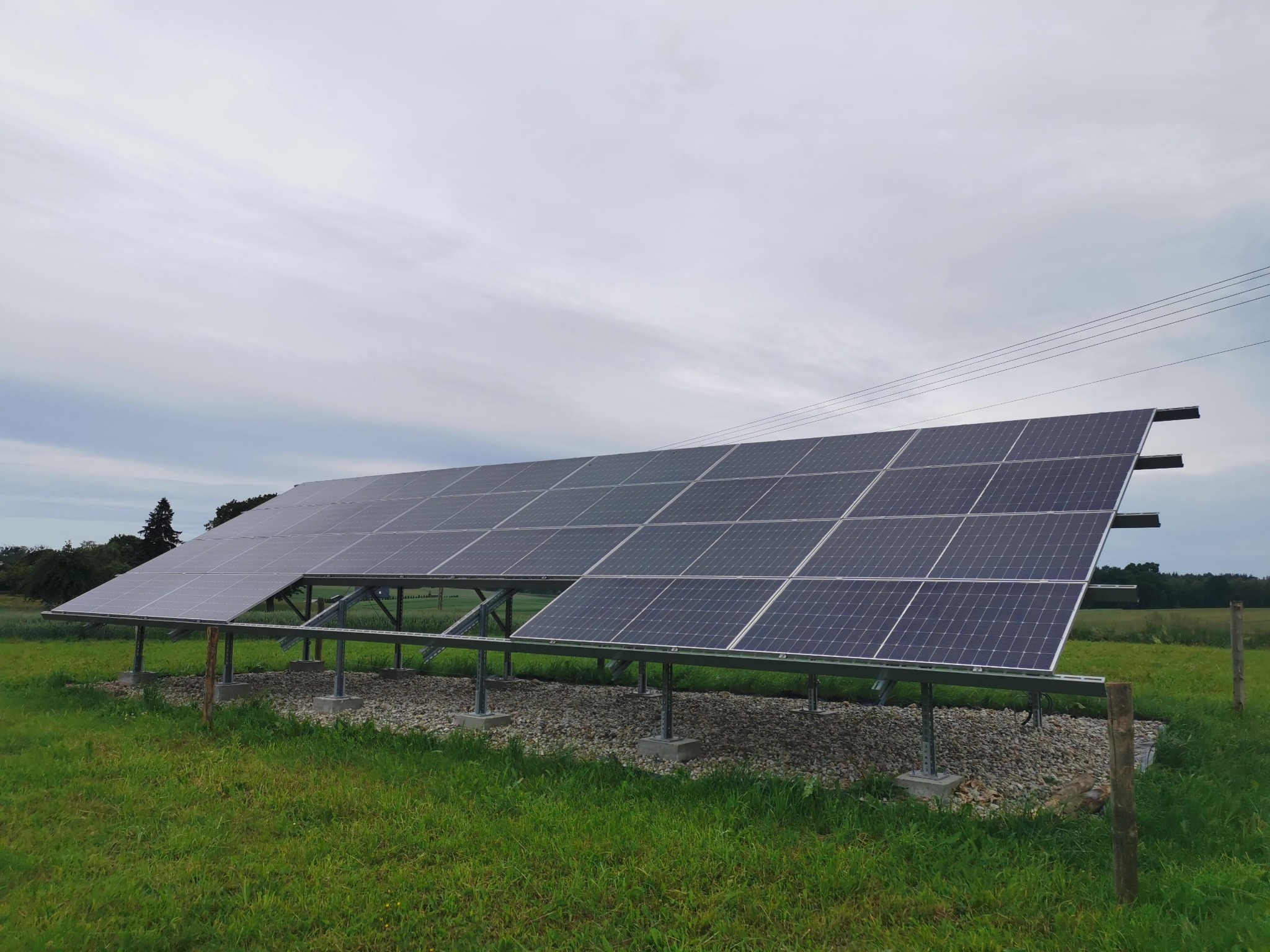 Rząd paneli słonecznych zamontowany na metalowej konstrukcji na łące, widoczne betonowe podstawy i kamienny podsyp, pochmurne niebo w tle.
