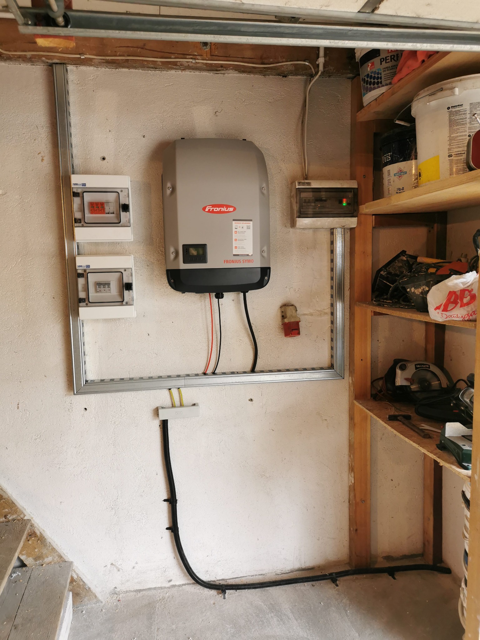 Szary falownik Fronius Symo zamontowany na ścianie w garażu, widoczne skrzynki elektryczne, metalowa rama montażowa, kable i fragment regału z narzędziami.
