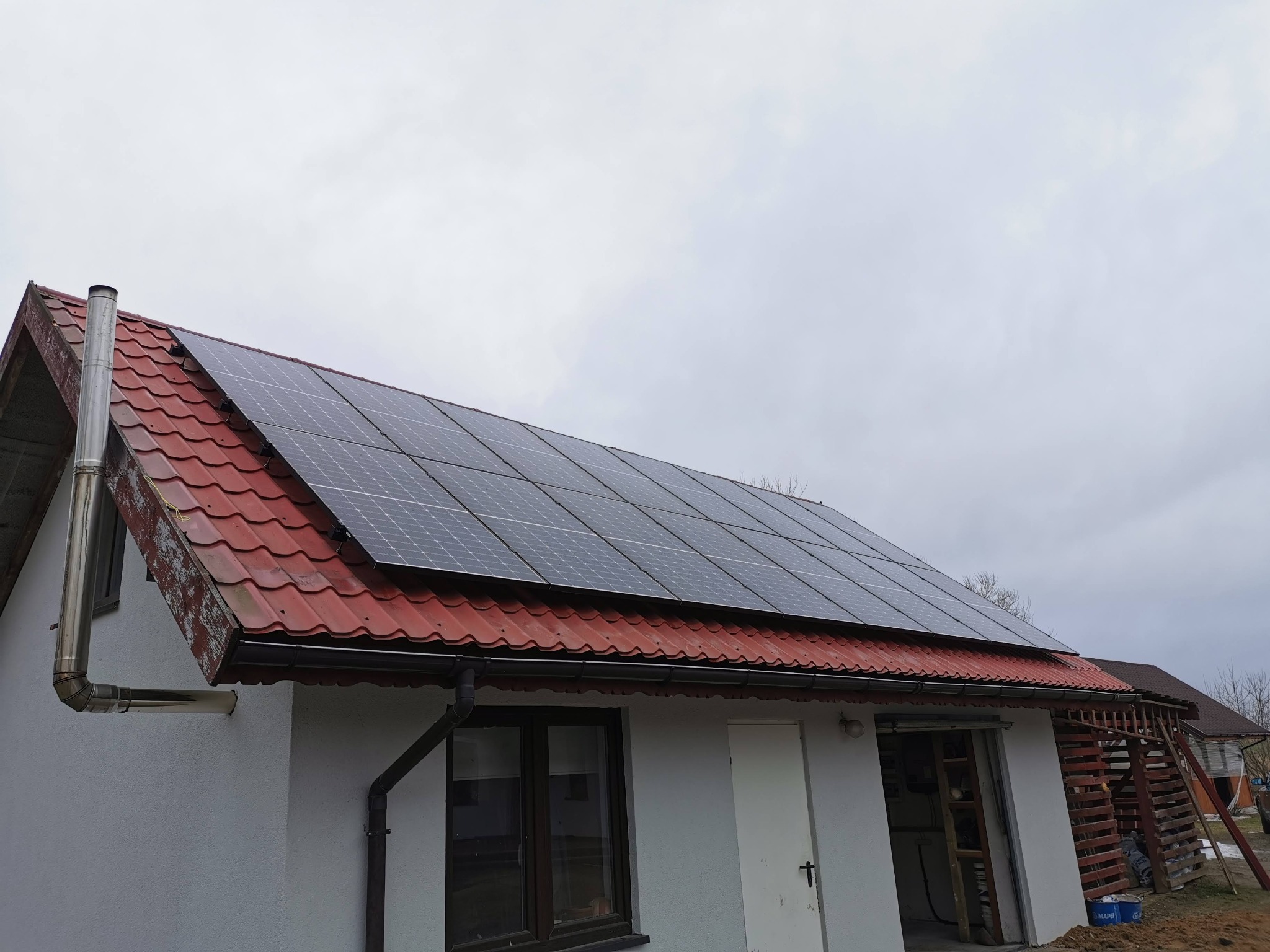 Dom jednorodzinny z panelami słonecznymi na czerwonym dachu, widocznym kominem i fragmentem garażu z drewnianą konstrukcją wsporczą, pochmurne niebo.