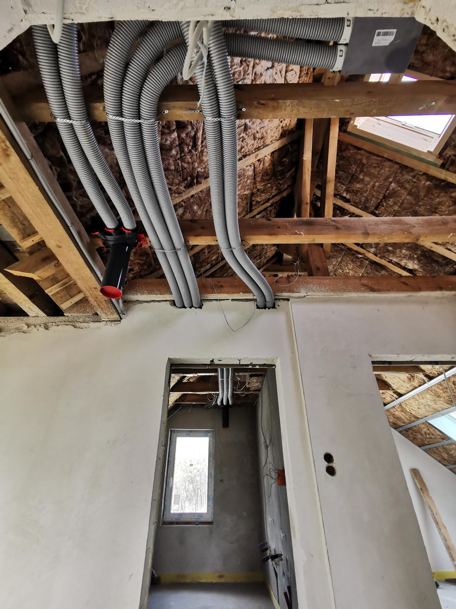 Szare, karbowane rury wentylacyjne prowadzone pod drewnianym stropem na poddaszu, z widoczną izolacją termiczną, połączone z czarną rozdzielnicą z czerwonymi elementami, widok z dołu na surową...