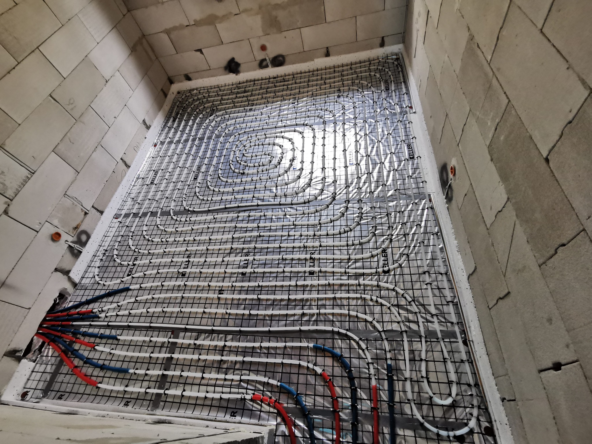 Instalacja ogrzewania podłogowego w surowym wnętrzu z bloczków, widoczne białe rury układane na folii aluminiowej i czarnej siatce, z czerwono-niebieskimi rurami doprowadzającymi.