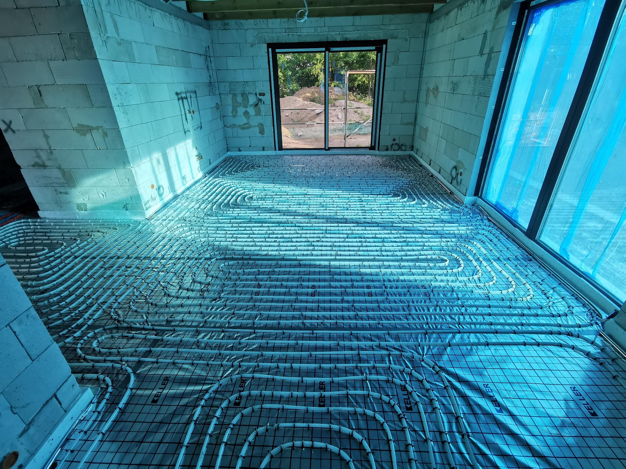 Rozłożone rury ogrzewania podłogowego na folii z siatką w surowym stanie, ściany z białych bloczków, widok na zewnątrz przez przeszklone drzwi balkonowe.