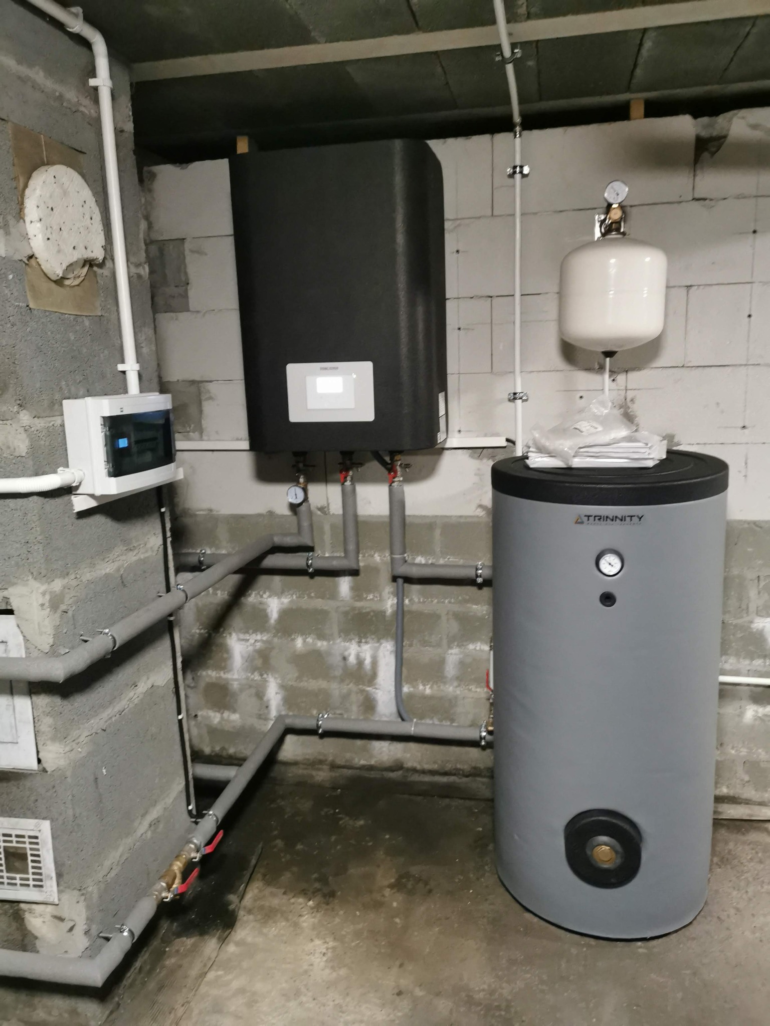 Szara instalacja z czarnym panelem sterowania oraz białym zbiornikiem wyrównawczym w surowym pomieszczeniu z betonowych bloczków.