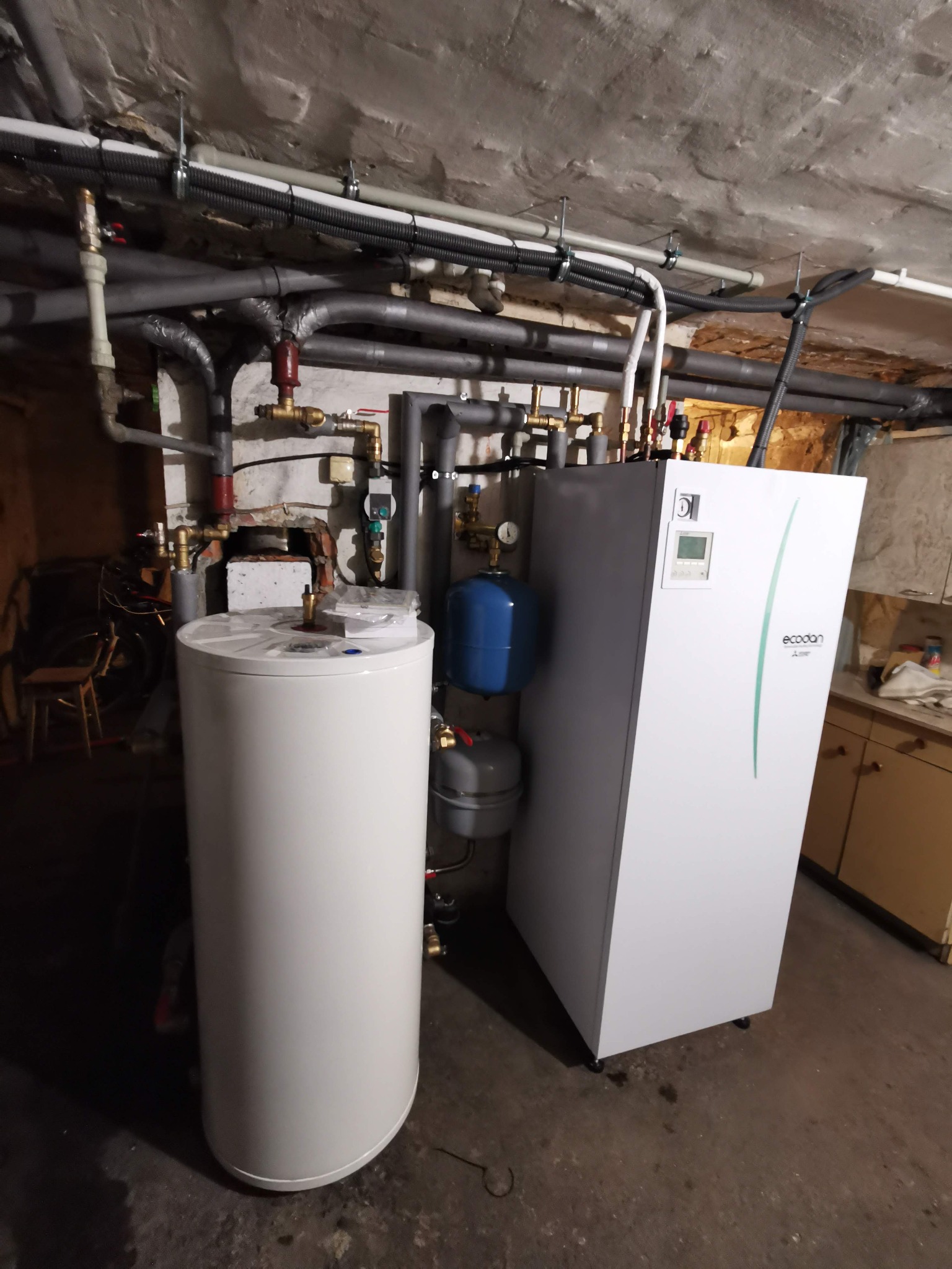 Biały zbiornik ciepłej wody użytkowej i jednostka zewnętrzna pompy ciepła Ecodan Mitsubishi w piwnicy, widoczne rury instalacyjne pod sufitem.