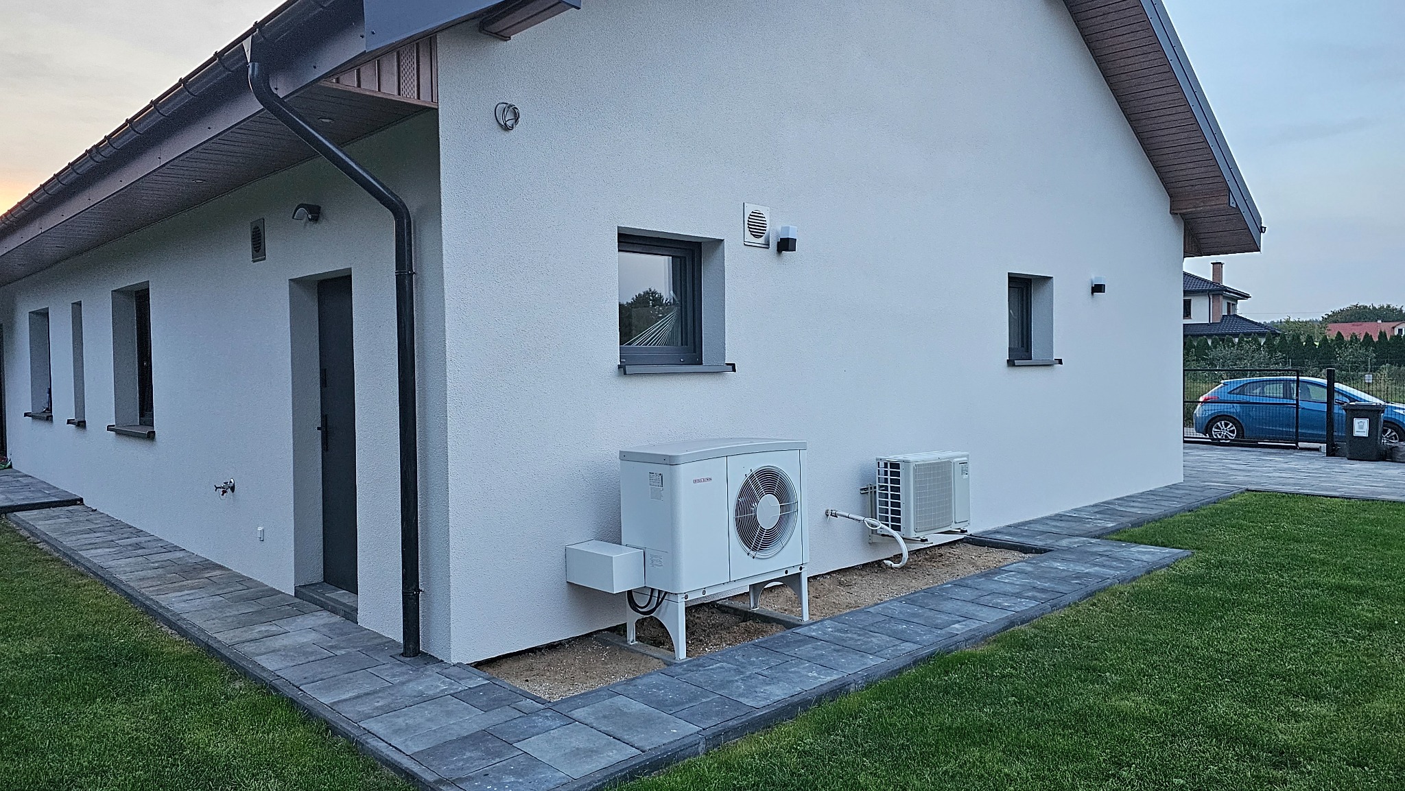 Biały dom jednorodzinny z zainstalowaną zewnętrzną jednostką pompy ciepła i klimatyzatora, widoczne okna, chodnik z kostki brukowej i trawnik.