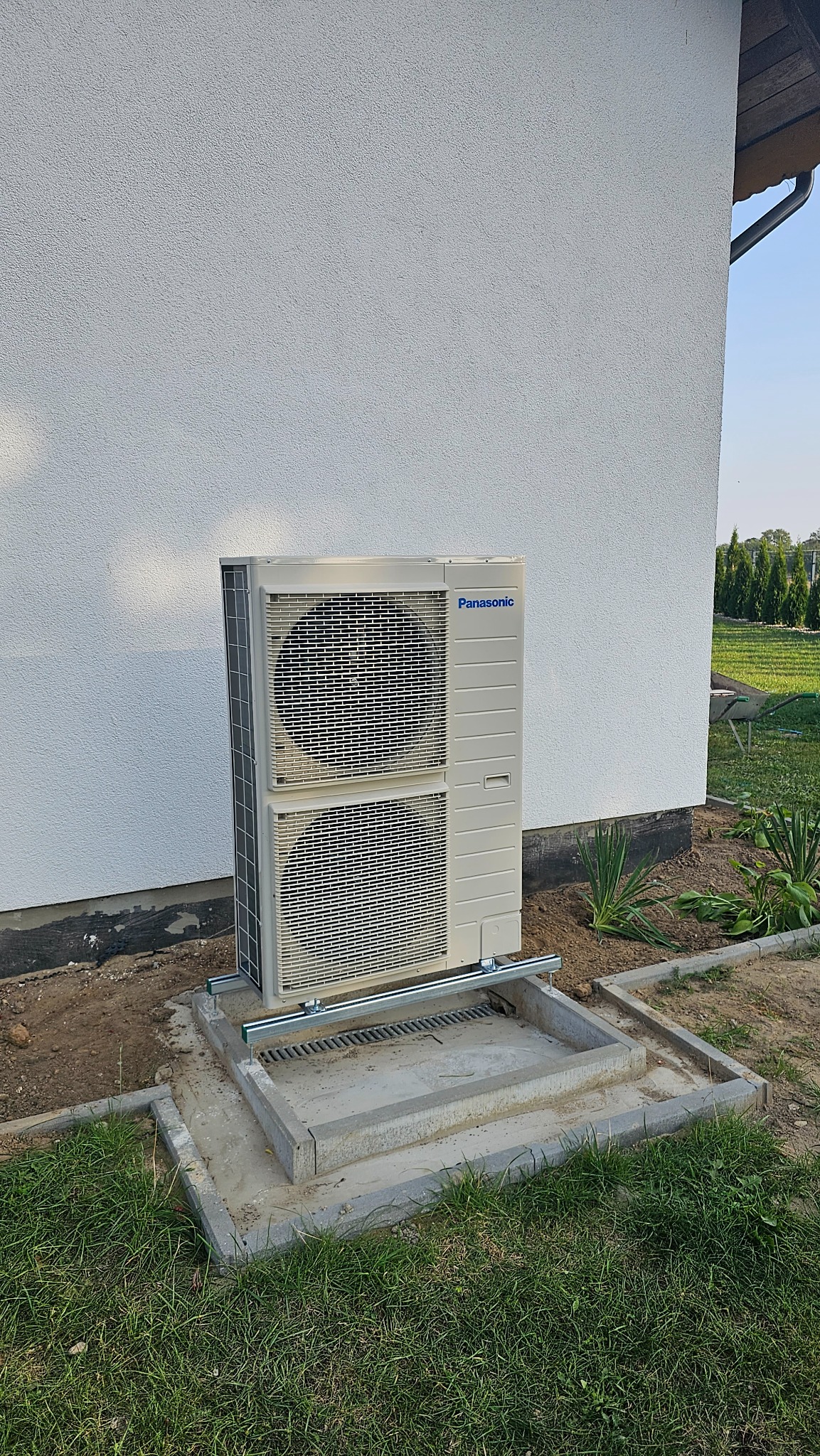 Zewnętrzna jednostka pompy ciepła Panasonic zamontowana na betonowej podstawie z odpływem, przy ścianie budynku z fakturą baranka, widok z boku w słoneczny dzień.