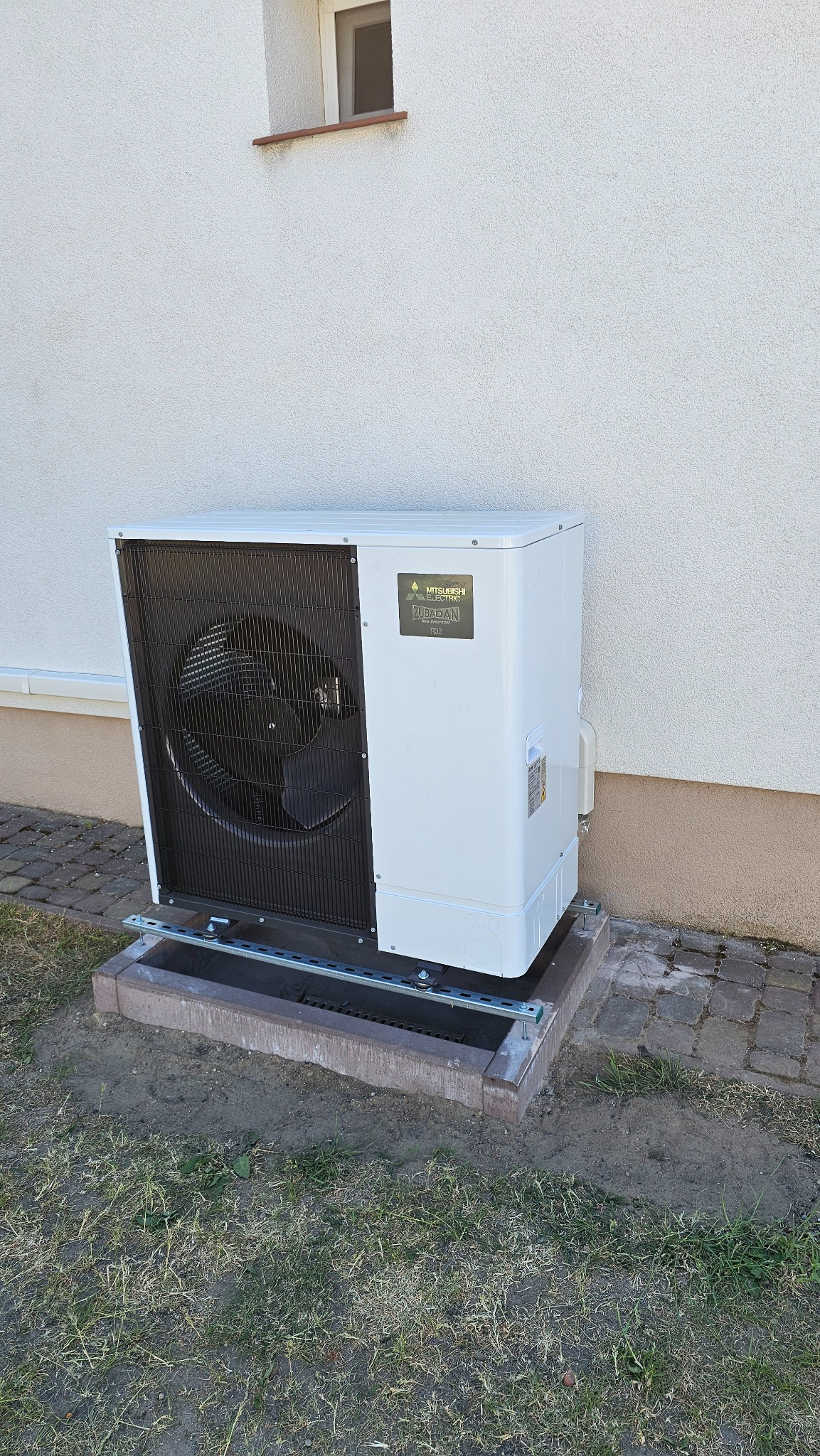 Biała pompa ciepła Mitsubishi Zubadan z czarnym wentylatorem, zamontowana na betonowej podstawie przy ścianie budynku z małym oknem.