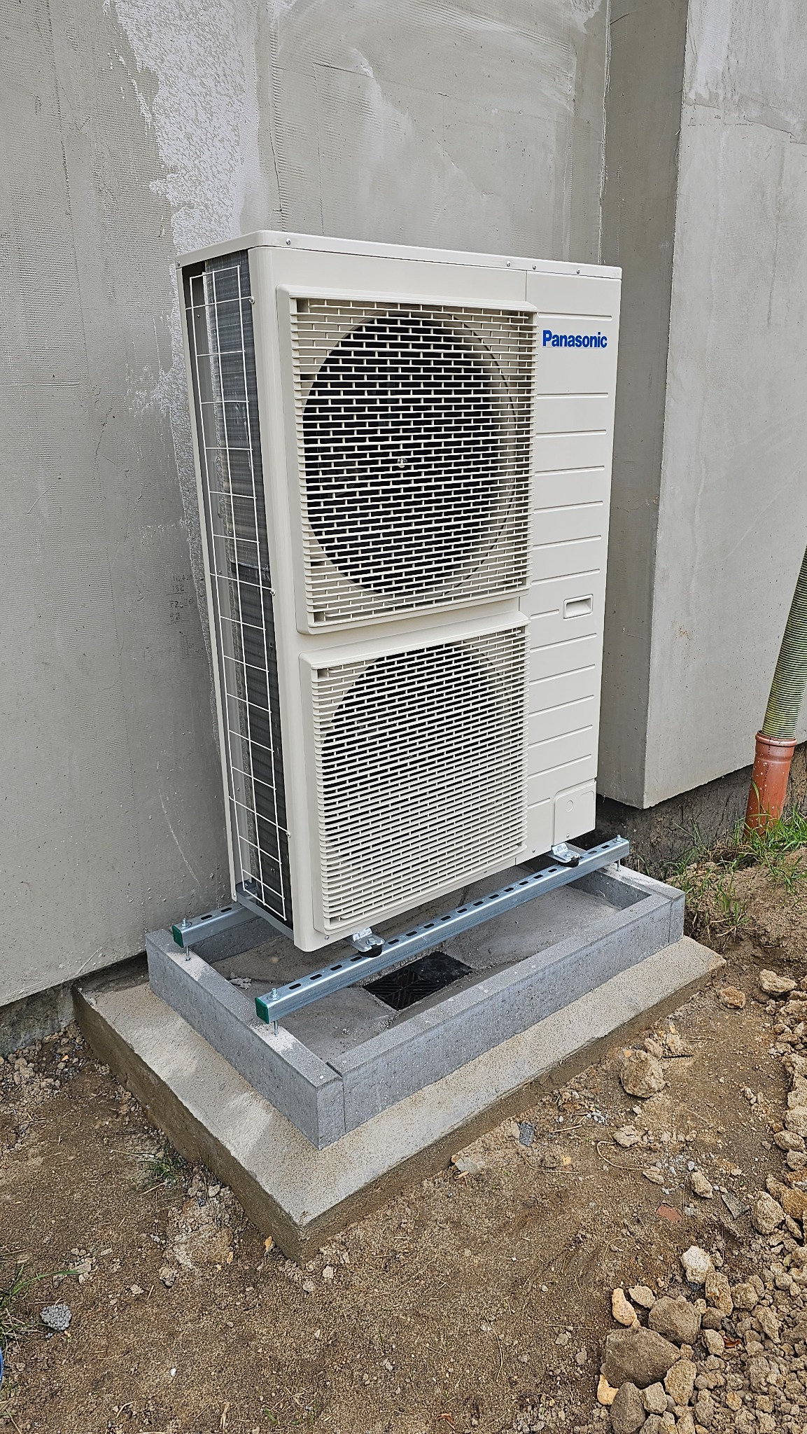 Biała jednostka zewnętrzna Panasonic, zamontowana na betonowej podstawie z szarą obudową i stalową konstrukcją wsporczą, przy szarej, surowej ścianie budynku.