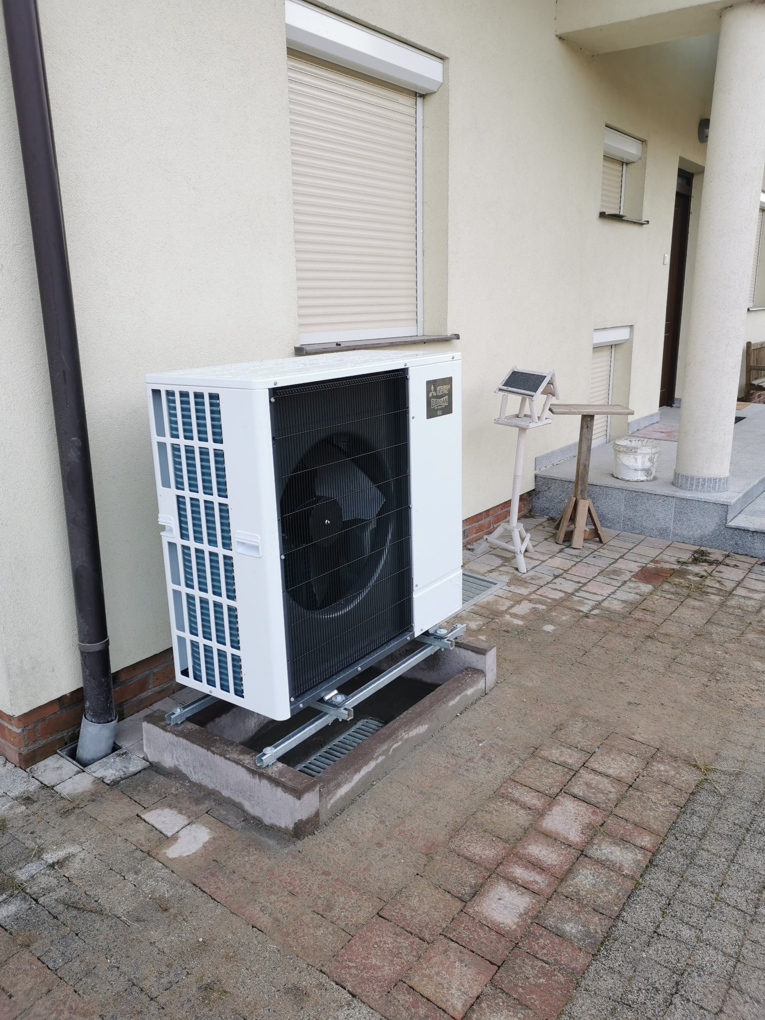 Biała jednostka zewnętrzna urządzenia grzewczego Mitsubishi Zubadan zamontowana na metalowej konstrukcji przy ścianie budynku z beżową elewacją i roletami zewnętrznymi.