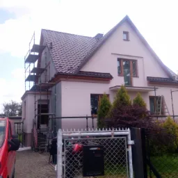 Dom jednorodzinny w trakcie prac elewacyjnych, widoczne rusztowania, częściowo ocieplona ściana, czerwony samochód dostawczy zaparkowany przed posesją.