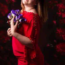 Kobieta w czerwonej sukience z odkrytymi ramionami trzyma bukiet fioletowo-białych róż na tle ciemnego, kwiatowego wzoru.