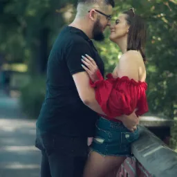 Para zakochanych, mężczyzna w czarnej koszulce i okularach obejmuje kobietę w czerwonej bluzce i dżinsowych szortach, stoją blisko siebie, dotykając się nosami na tle zieleni.