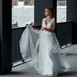 Panna młoda w koronkowej sukni z tiulowym dołem, trzymająca delikatnie materiał, na tle nowoczesnej architektury z widokiem na drzewa.