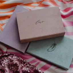 Trzy welurowe pudełka w pastelowych kolorach (liliowy, pudrowy róż i miętowy) ułożone na wzorzystej tkaninie, z napisem 'love' na różowym i rysunkiem piórka na miętowym, obok fragmentu błyszczącej...