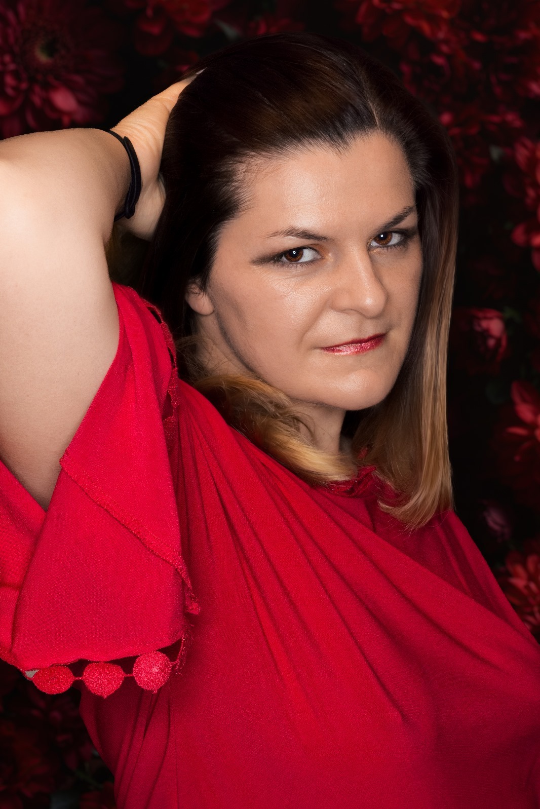 Portret kobiety w czerwonej sukience na tle ciemnych kwiatów, włosy z pasemkiem blondu opadają na ramię.