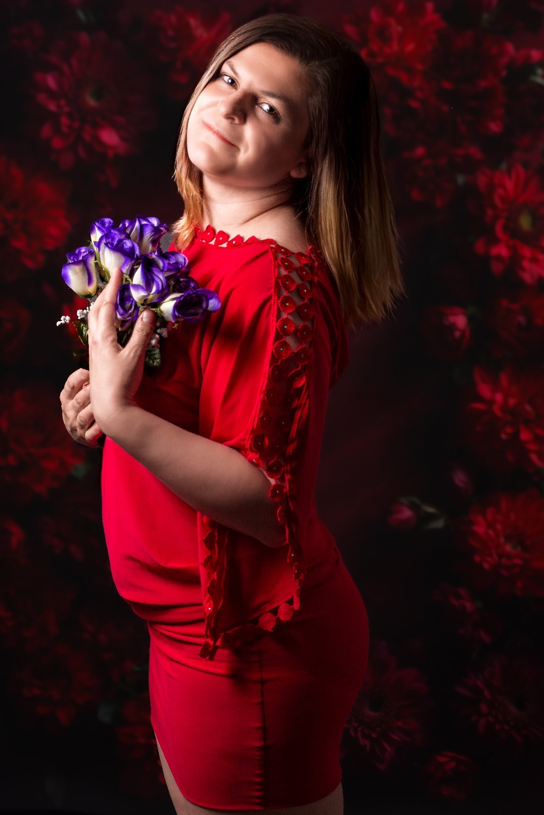Kobieta w czerwonej sukience z odkrytymi ramionami trzyma bukiet fioletowo-białych róż na tle ciemnego, kwiatowego wzoru.