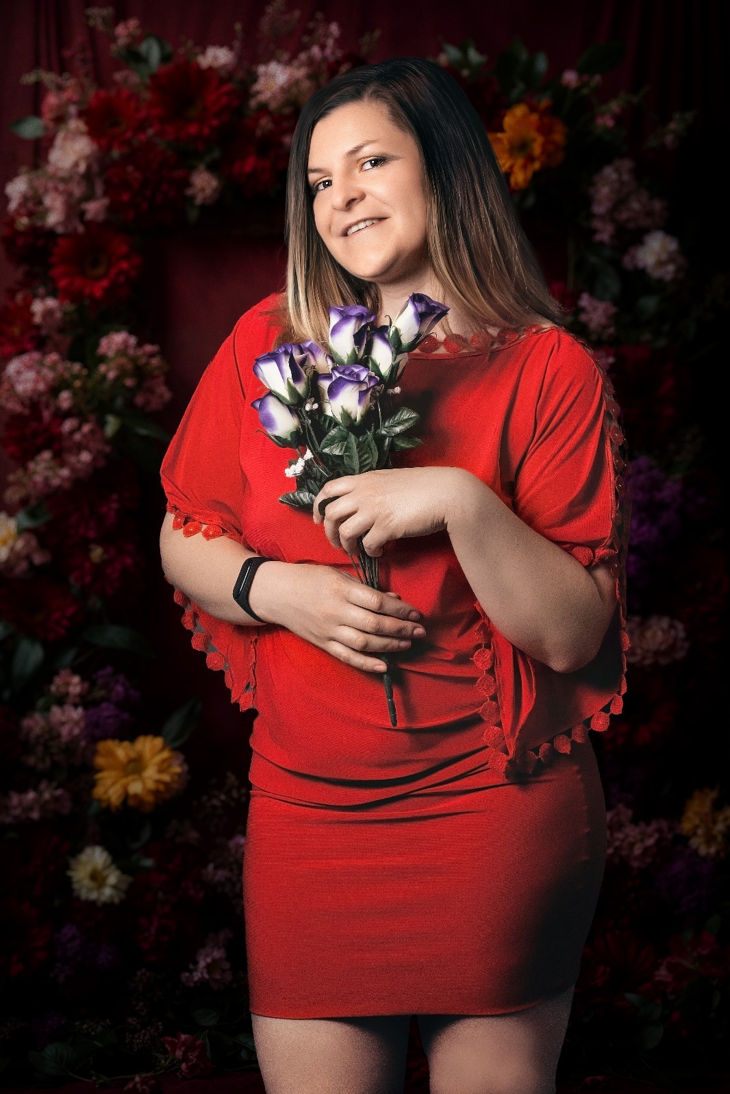 Uśmiechnięta kobieta w czerwonej sukience trzyma bukiet białych róż z fioletowymi końcówkami na tle ciemnego tła z kwiatami.