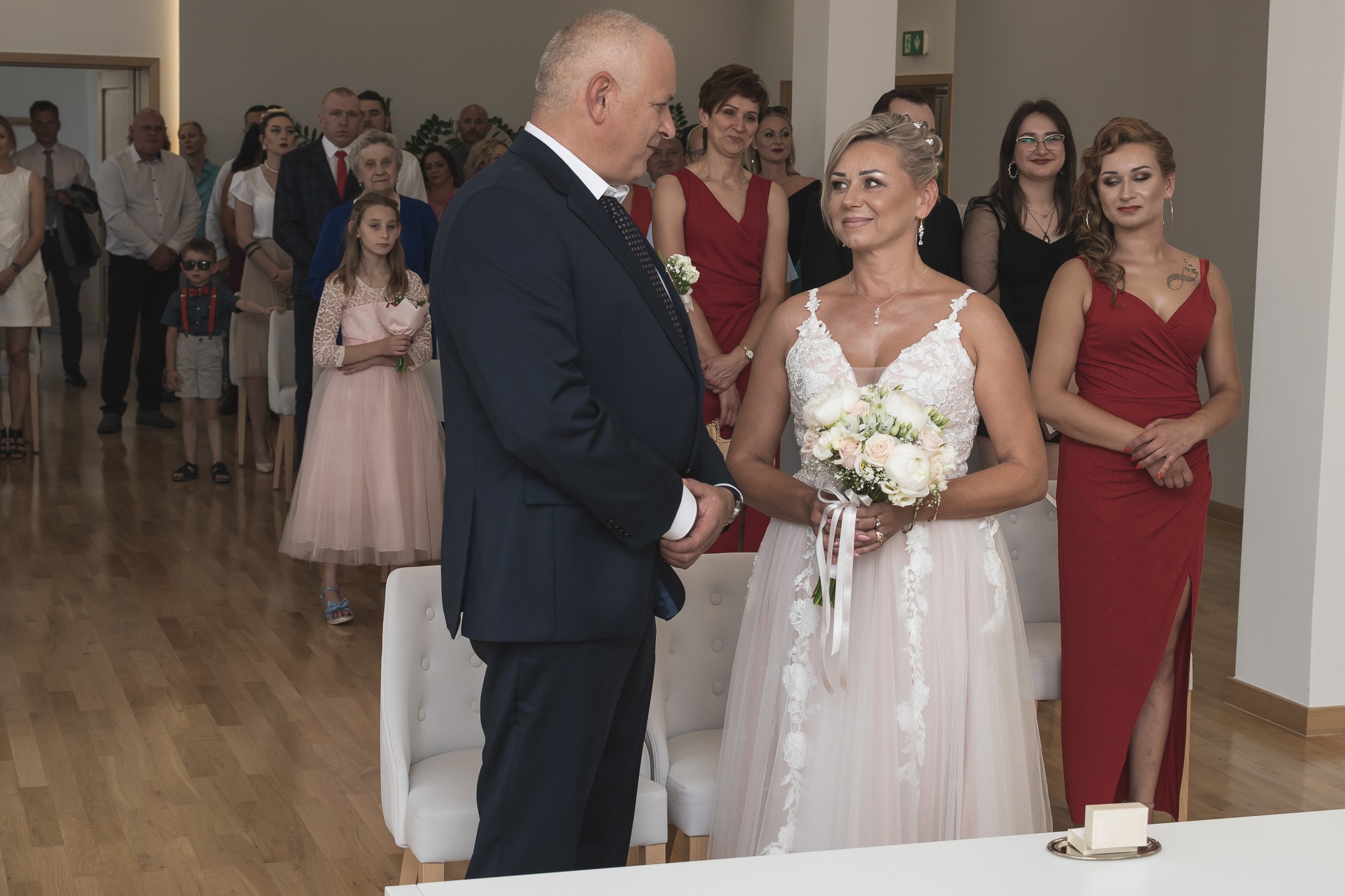 Panna młoda w koronkowej sukni ślubnej trzyma bukiet białych róż, obok stoi pan młody w granatowym garniturze, w tle goście weselni.