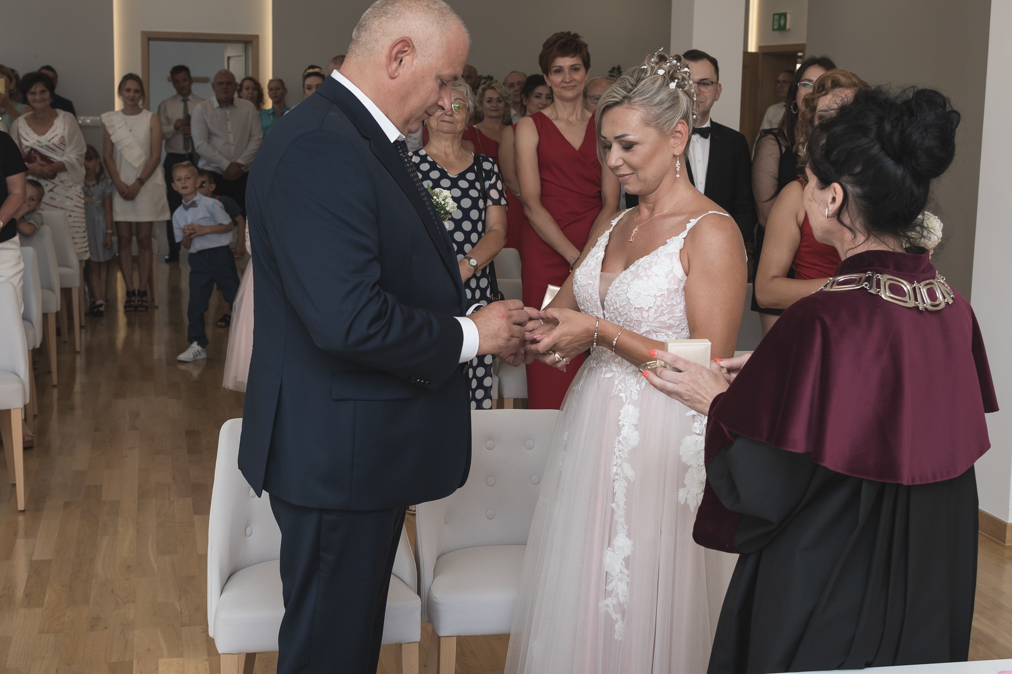 Moment zakładania obrączki podczas ceremonii ślubnej w jasnym wnętrzu, z widocznymi gośćmi w tle.