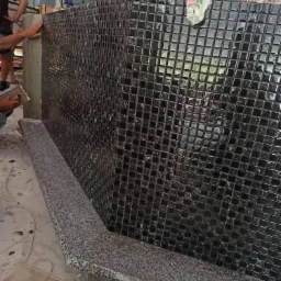 Proces układania czarnej mozaiki kwadratowej na ścianie, z widocznym fragmentem szarego, granitowego blatu i narzędziem glazurniczym w ręku wykonawcy.