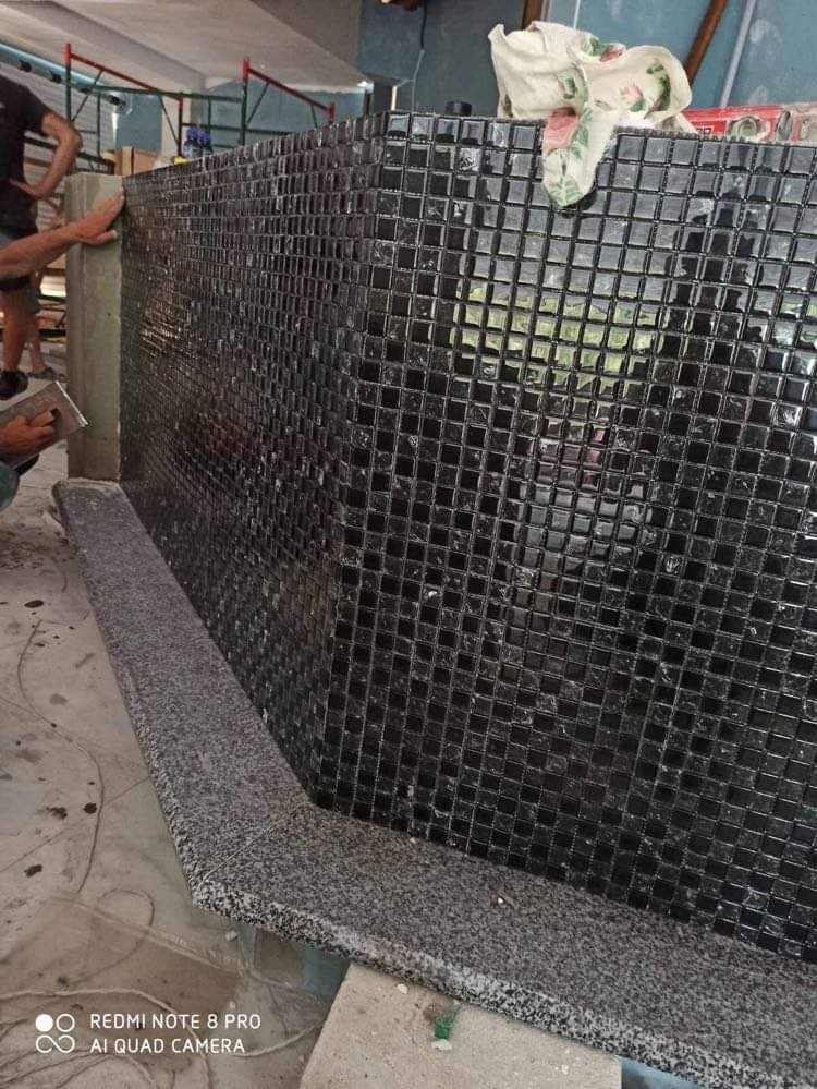 Proces układania czarnej mozaiki kwadratowej na ścianie, z widocznym fragmentem szarego, granitowego blatu i narzędziem glazurniczym w ręku wykonawcy.