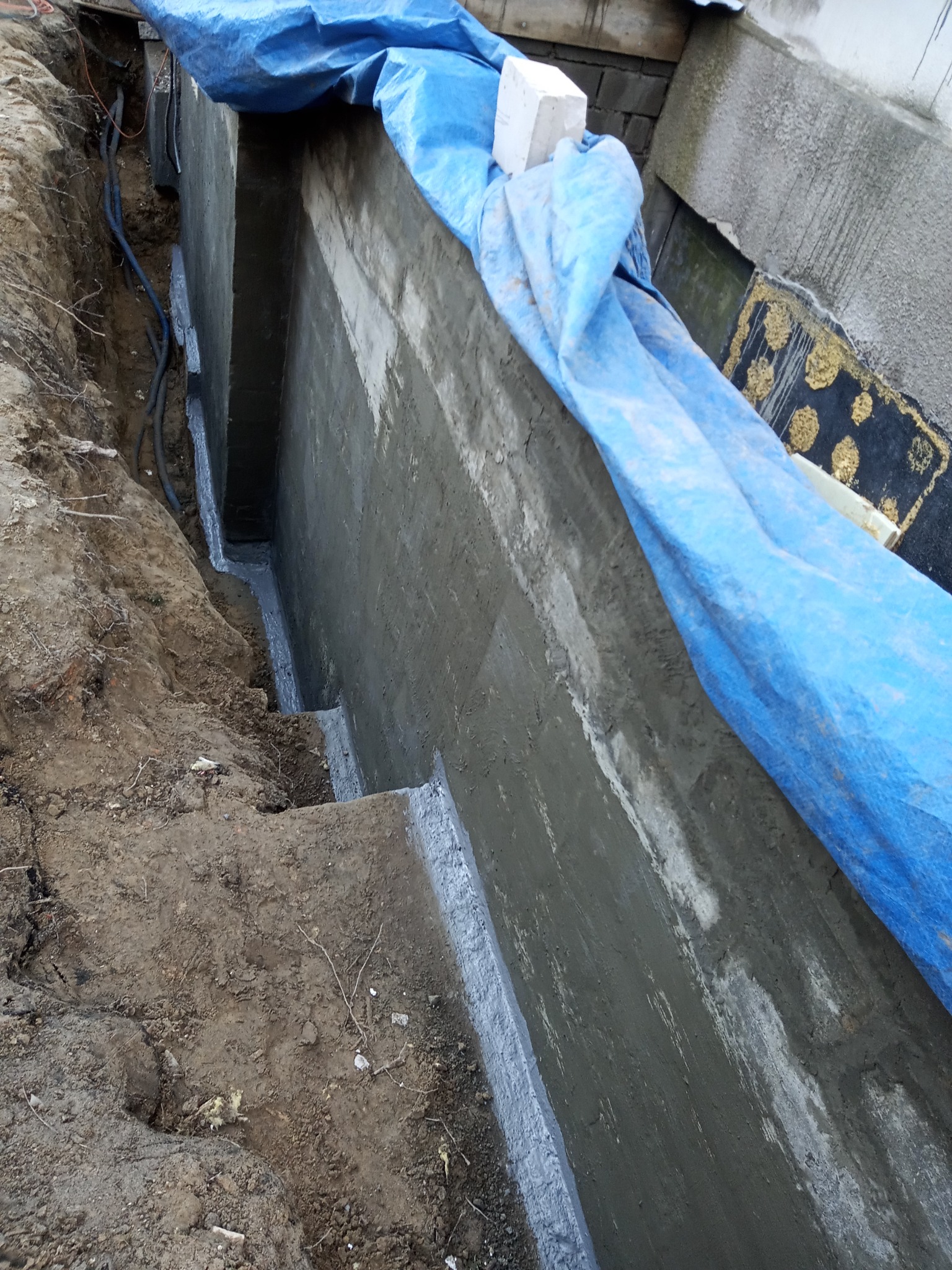 Wykonanie izolacji fundamentu z widoczną warstwą hydroizolacji, zabezpieczone niebieską folią budowlaną, z odkrytymi kablami w wykopie.