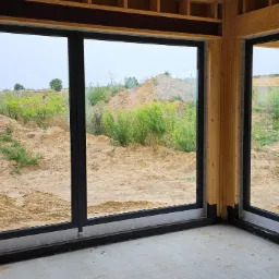 Surowy stan budowy: Duże, czarne ramy okienne z widokiem na piaszczysty teren z roślinnością, widoczne elementy konstrukcyjne budynku.
